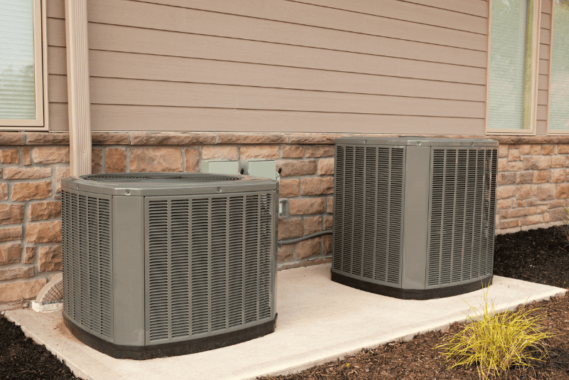 Two outdoor air conditioning units sit on a concrete pad next to the exterior wall of a building with siding and brick accents.