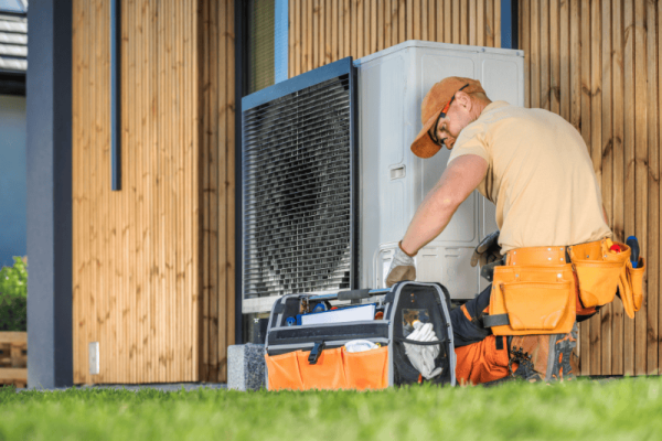 A technician in a tan uniform and orange cap services an outdoor HVAC unit, with a tool bag open on the grass beside him.
