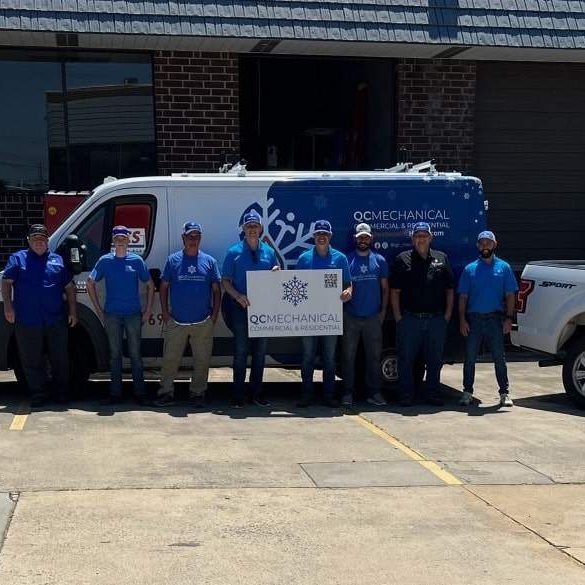 A group of eight people in matching blue shirts stands in front of a van and two trucks, all labeled "OG Mechanical Services," outside a building on a sunny day.