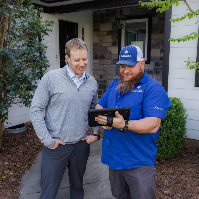 Two men stand outside a house, looking at a tablet held by the man in a blue shirt and cap. The man in a gray sweater smiles as he observes the tablet. Shrubs and trees are visible around them.