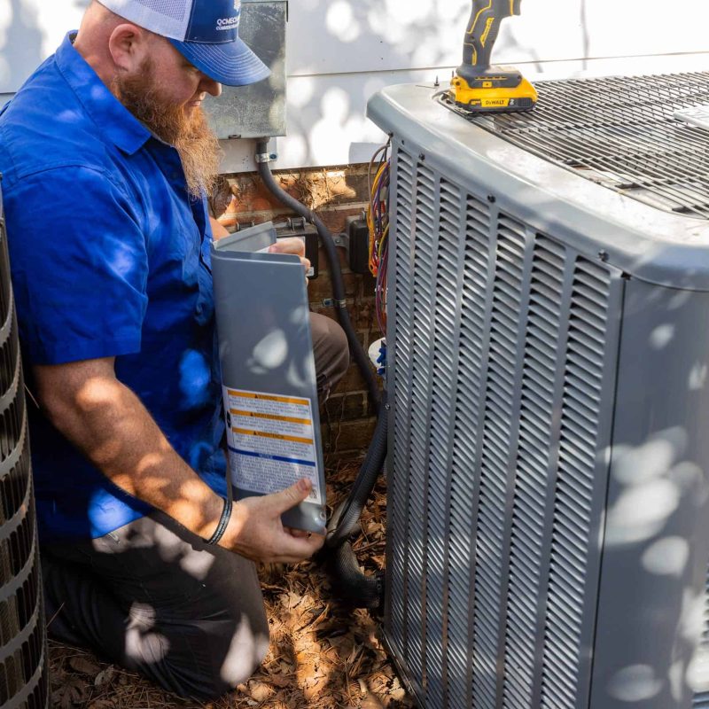 Technician in a blue shirt and cap works on an outdoor air conditioning unit, holding a part while a yellow drill rests on top of the unit.