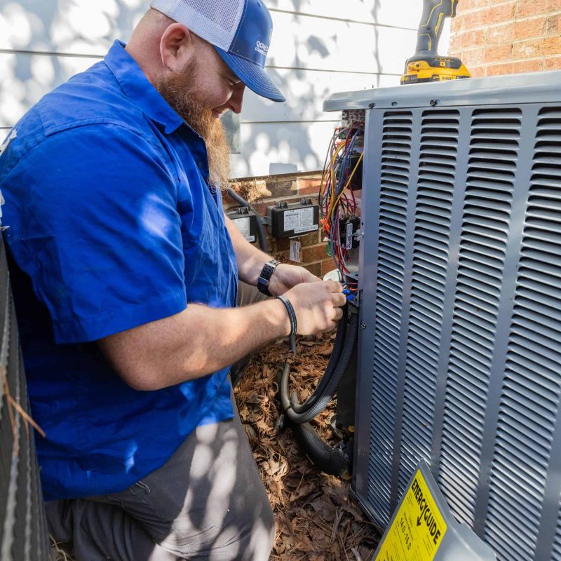 A technician in a blue shirt works on an air conditioning unit outdoors. A yellow drill is placed on top of the unit.