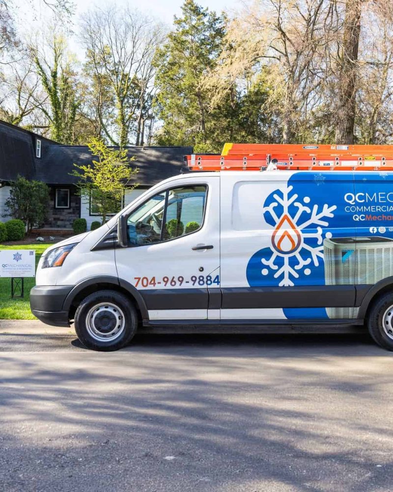 A white QC Mechanical service van, with a snowflake and flame logo, is parked on a residential street in front of a house. The van's doors display contact information and company branding.