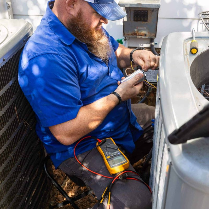 A bearded technician in a blue shirt and cap uses a multimeter to check electrical connections on an HVAC unit outside a white building.