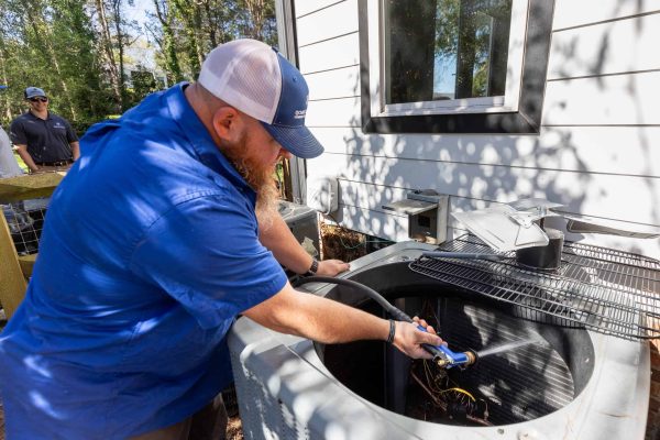 A man in a blue shirt and white cap works on an outdoor air conditioning unit. Another man stands in the background, partially visible.