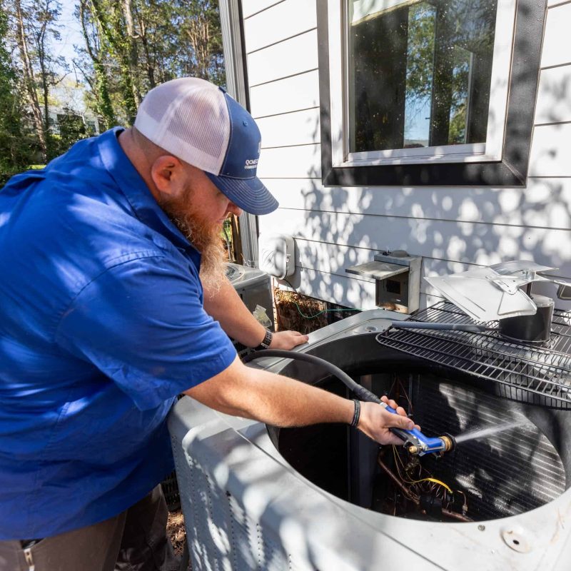 A man in a blue shirt and white cap works on an outdoor air conditioning unit. Another man stands in the background, partially visible.