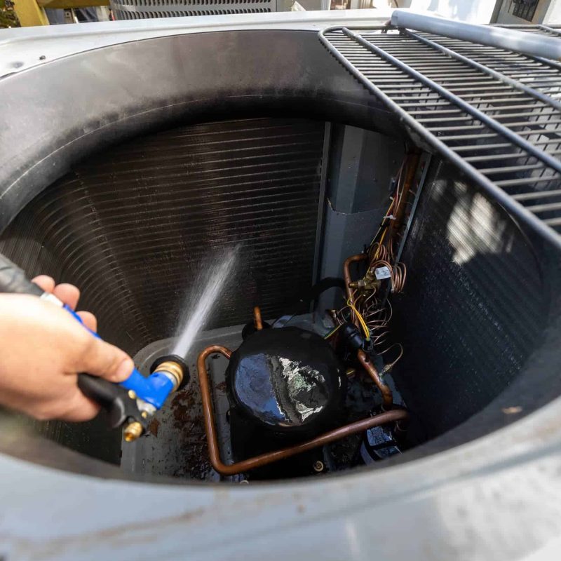 A person is using a hose to clean the inside of an air conditioning unit, focusing on the coils and components.