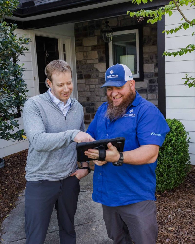 Two men standing outside a house, one wearing a blue shirt and cap holding a tablet, and the other in a gray sweater and dress shirt looking at the tablet.