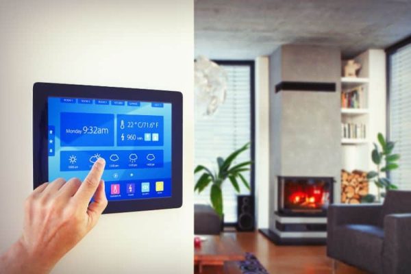 A hand interacts with a smart home panel displaying the time, weather, and control icons. In the background, a modern living room with a fireplace and a large plant is visible.