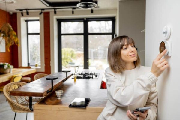 A person adjusts a thermostat on a wall in a modern kitchen. They are holding a smartphone and wearing a light-colored sweater.