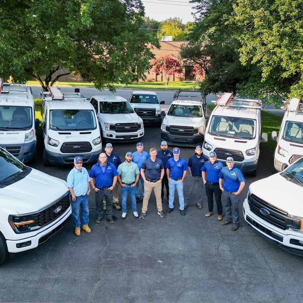 A group of eight men dressed in casual and work clothes stands in front of a fleet of white utility trucks and vans parked outdoors, surrounded by trees and greenery.