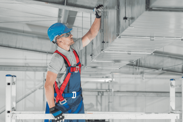 A worker wearing safety gear, including a blue helmet and harness, inspects or installs a metal duct on a raised platform inside an industrial building.